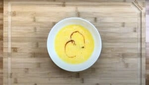 Creamy beaten eggs topped with a drizzle of sauce in a white bowl on a wooden board.